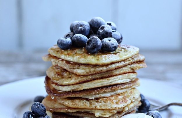 plate with a stack of about 10 pancakes on top of each other with a bunch of blueberries sitting on top of the pancake stack