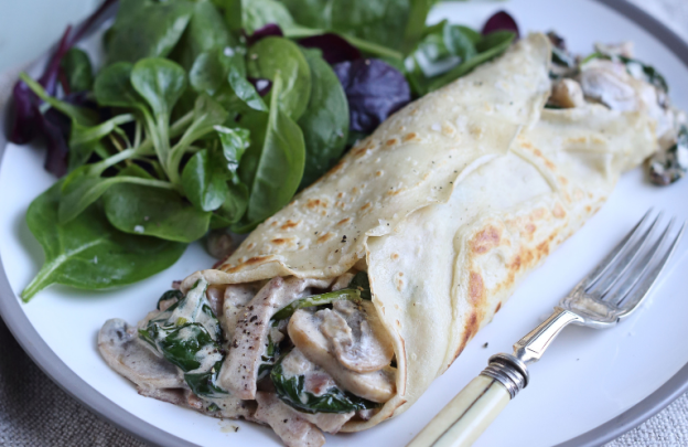 Plate with fork at the front and then a pancake rolled up filled with mushrooms and then a bunch of green leaves beside the pancake