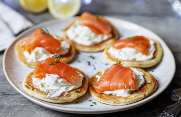 Plate with small pancakes on the plate in a circle, with cream cheese on top of each mini pancake and then topped with smoked salmon