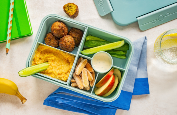 Lunchbox in centre of image with falafel in one corner, cucumber slices, hummus and nuts in the lunch box too.