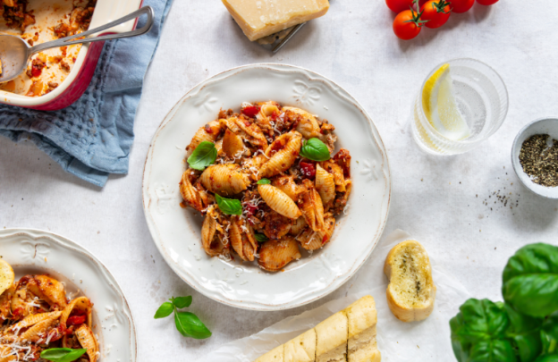 white bowl in centre of image with fresh pasta and bolognese with a glass of water to the right hand side and garlic bread at the bottom of the image.