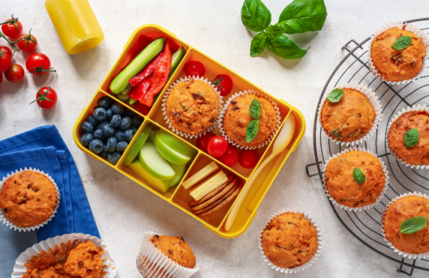 Lunch box in centre of image with savoury muffins in the middle with blueberries, apple slices, crackers and cumber slices in lunchbox too.